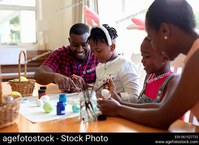 African american parents with son and daughter wearing bunny ears painting colourful eggs