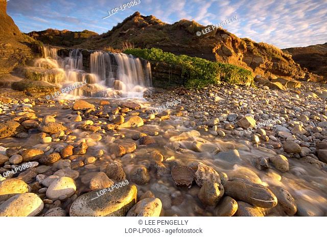 England, Dorset, Osmington Mills, A waterfall flowing down over a pebbly beach on the jurassic coast