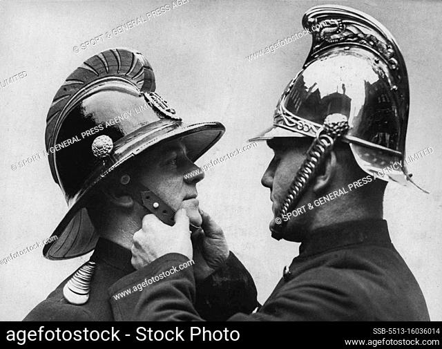 New Helmet for Firemen. A fireman wearing the old type helmet adjusts the strap of a new model. The seining brass helmet of the London fire Brigade