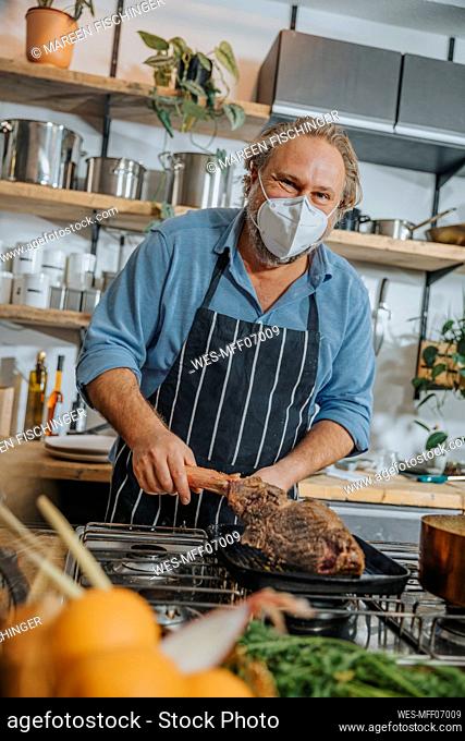 Mature expertise wearing face mask grilling tomahawk steak in frying pan while standing in kitchen