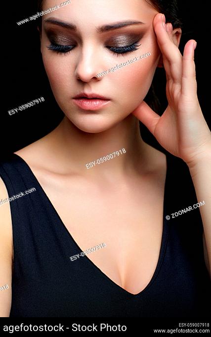 Beauty portrait of young woman with hand near face on black background. Brunette girl with evening female makeup and black dess touches face with fingers