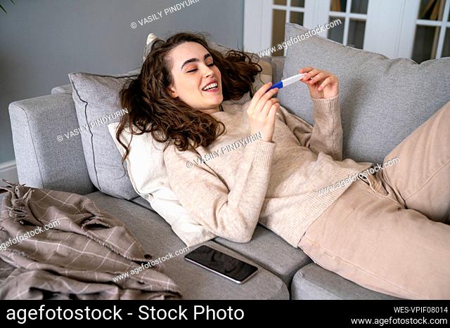 Happy young woman looking at pregnancy testing kit relaxing on sofa