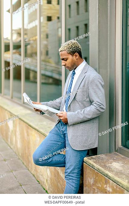 Businessman leaning against wall, reading newspaper