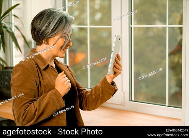 Pretty mature woman communicates online on digital tablet standing near window. Smiling woman connecting with friends by internet