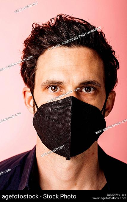 Businessman wearing black color protective face mask staring while standing against colored background