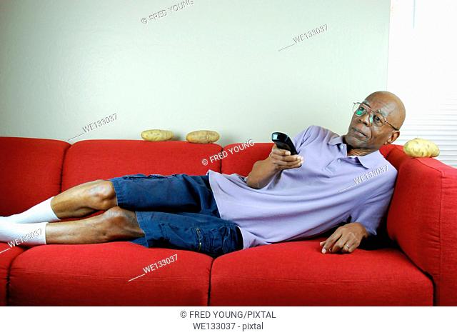 A senior citizen relaxing on a couch with a remote control in hand and potatoes on the couch