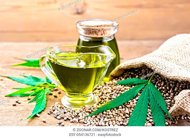 Hemp oil in a glass sauceboat and a jar with grain in a bag, leaves and stalks of cannabis on a old wooden board background