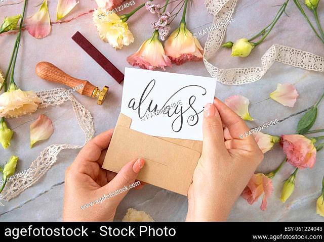 Hands with Card ALWAYS inside envelope near by pink flowers, wax seal and ribbons on a marble table top view. Romantic scene with handwritten card flat lay