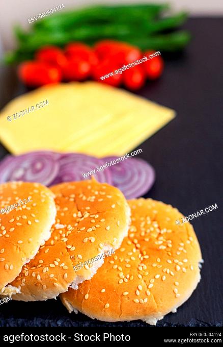 Preparation for the preparation of a hamburger, all ingredients: cheese, salad, tomato, onion, bun lie on a black stone background. Close-up