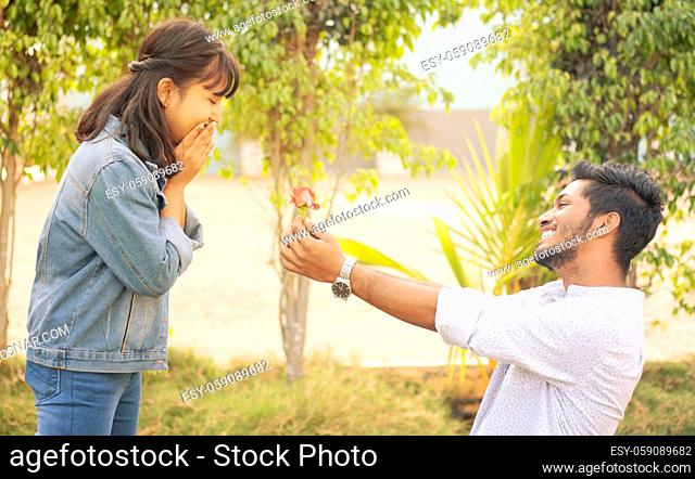 Concept of teenage love or affection - young man proposing to smiling excited girlfriend standing on knee with red rose on valentines day