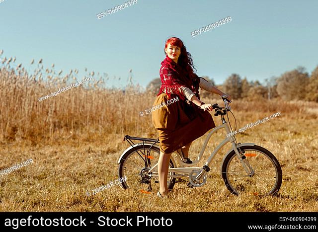 Beautiful young woman in long skirt and wool scarf riding retro style bicycle in field. Outside shot. Autumn season. Copy space
