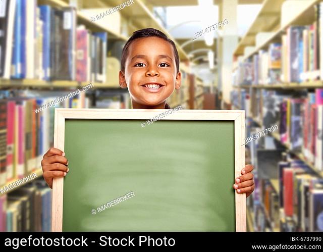 Happy smiling hispanic boy holding empty chalk board in library