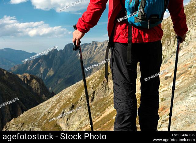 The body of man with a backpack and trekking poles stands on top of a rock against the background rocky valley high in the mountains