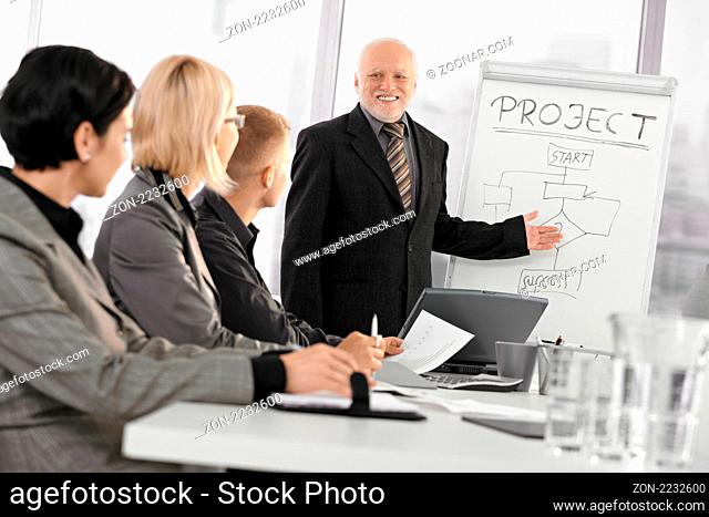Smiling senior businessman training project success to colleagues, pointing at whiteboard, audience listening