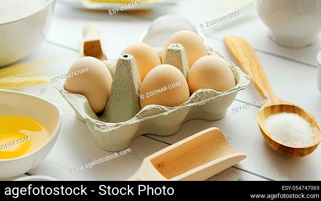 Closeup shot of various utensils and fresh ingredients for pasta preparation lying around carton with chicken eggs on white timber tabletop