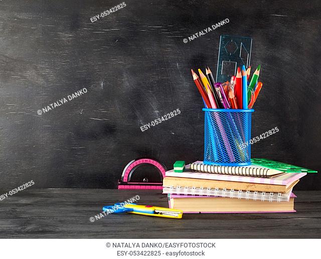 stack of notebooks, blue glass with multi-colored wooden pencils and pens, rulers and an eraser on the background of an empty black chalk board