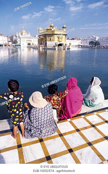 Women and children sitting on walkway beside sacred pool looking across to the Golden temple reflected in rippled surface of water