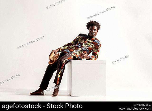 Barefooted african man uncomfortble sit on cube. Indoor, isolated on gray background