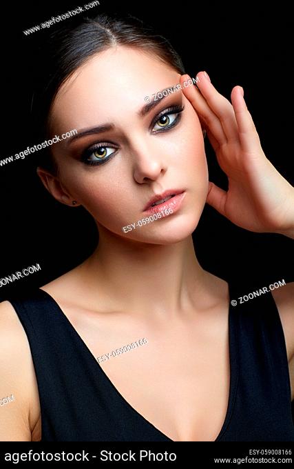 Beauty portrait of young woman with hand near face on black background. Brunette girl with evening female makeup and black dess touches face with fingers