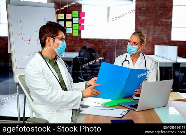 Diverse male and female doctor wearing face masks sitting in hospital office discussing paperwork