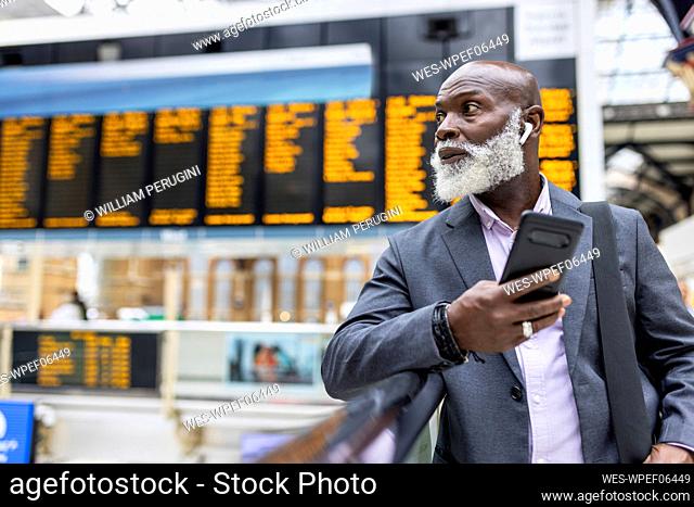 Senior businessman with smart phone listening to music at railroad station