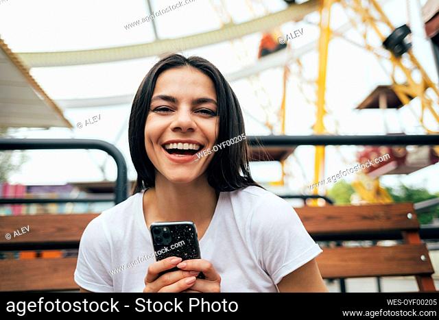 Woman holding smart phone laughing while sitting on bench at amusement park