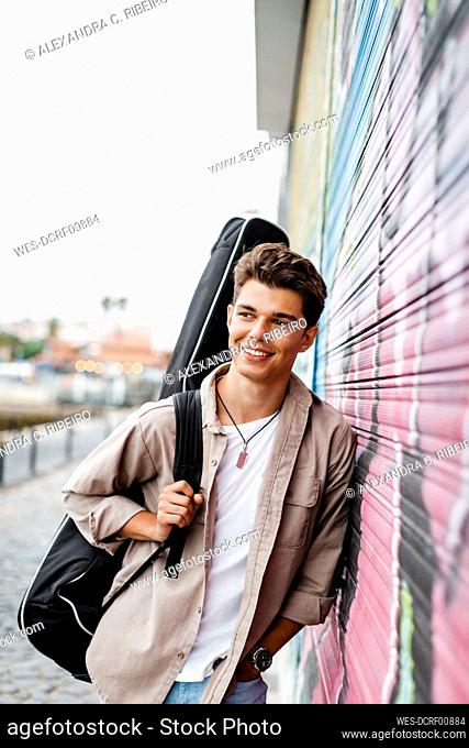 Smiling young man looking away while holding guitar and leaning on shutter with graffiti