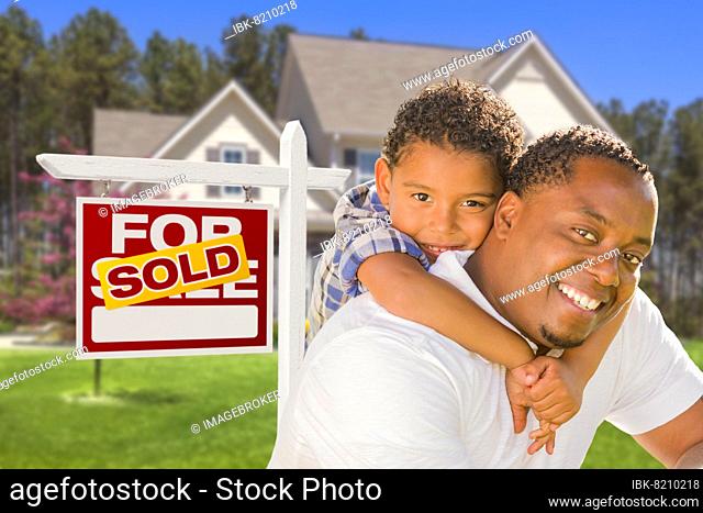 Happy mixed-race father and son in front of sold real estate sign and new house