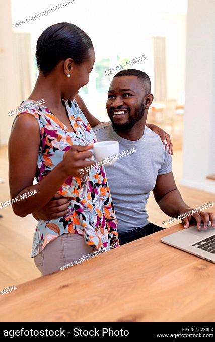 Happy african american couple embracing in kitchen, man using laptop woman holding coffee