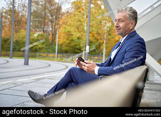 Senior male professional waiting while sitting with smart phone at bus stop