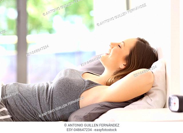 Side view portrait of a happy girl relaxing lying on a bed of an hotel room or apartment with a green background outdoors