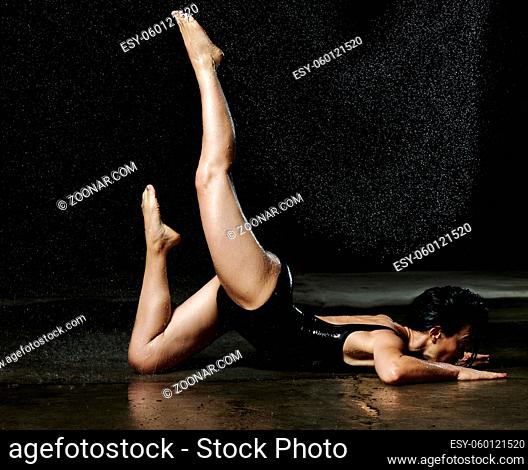 woman with black hair lies on the ground under raindrops on a black background. Woman dressed in black bodysuit, legs raised up