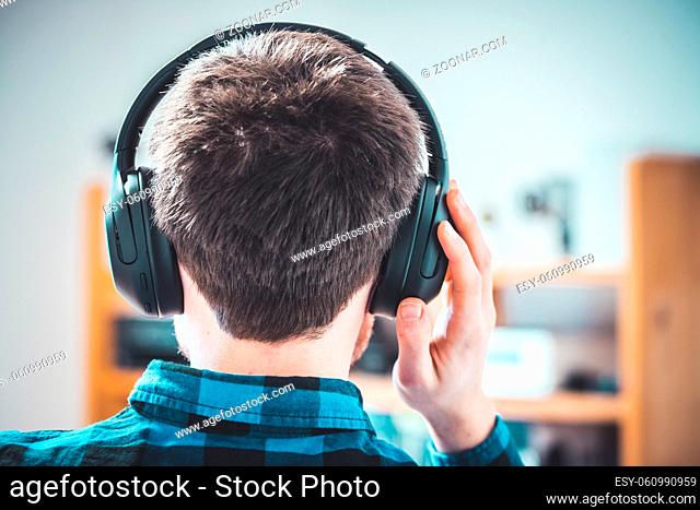 Young Caucasian man with checked blue shirt is listing to music with wireless headphones. Back of the head