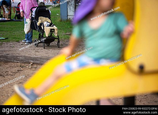 SYMBOL - 01 July 2023, Berlin: A toddler sits on the slide of a playground while an older adult holds a stroller in the background