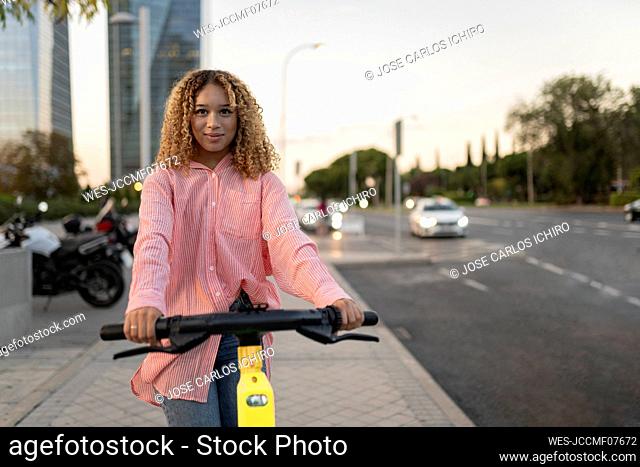 Young curly haired woman riding electric push scooter on footpath
