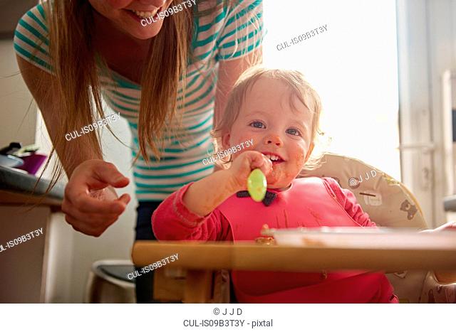 Toddler sitting in highchair, eating meal, mother standing behind her