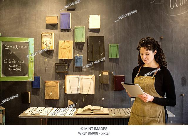Woman with curly brown hair wearing apron standing in her pottery shop, looking at digital tablet, display of books and jewellery