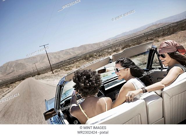 Three young people in a pale blue convertible car, driving on the open road across a flat dry plain