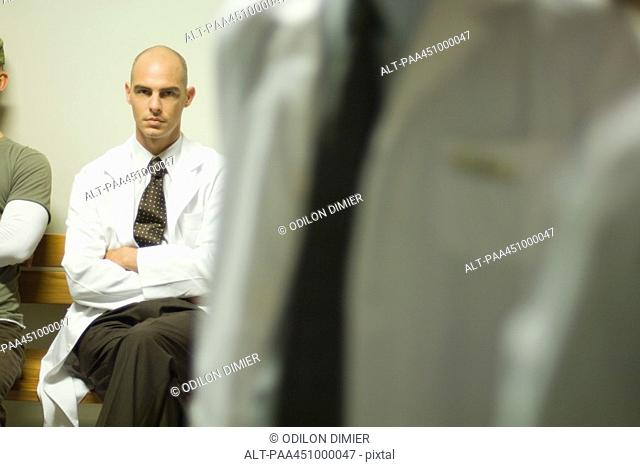 Male doctor sitting with arms folded, looking at camera, focus on background