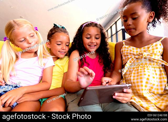 Smiling multiracial elementary schoolgirls using digital tablet while sitting on couch in school