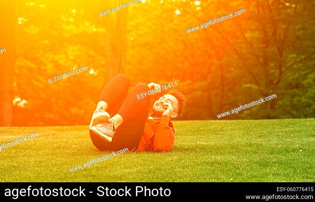 Sport man training in green park. Handsome man in red jacket lying on green grass and doing different exercises for abs. Toned