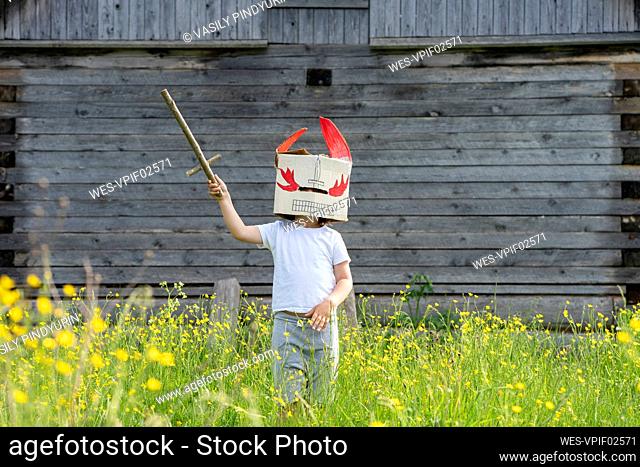 Boy wearing mask holding toy sword while standing amidst plants against cottage