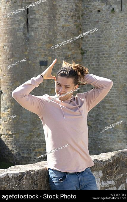 Young man in pink hoodie ties his hair in an exaggerated gesture that recalls the stylized mine of Kabuki theatre, Carcassonne Citadel; Occitanie; France