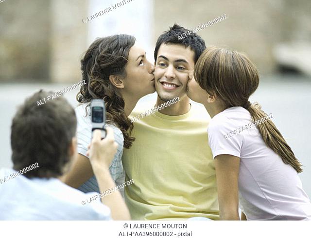 Teen girls kissing boy's cheeks while second boy takes photo