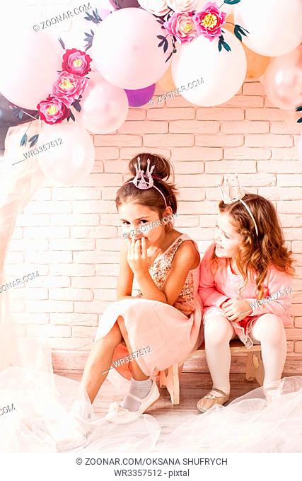 Two beautiful little girls with crowns under birthday balloon and paper flower arch decorations. Childish photozone for celebration