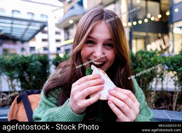 Happy young woman eating sandwich in front of building