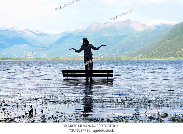 Woman standing on a bench in a flooding alpine lake maggiore, with snow-capped mountain in background in ticino switzerland