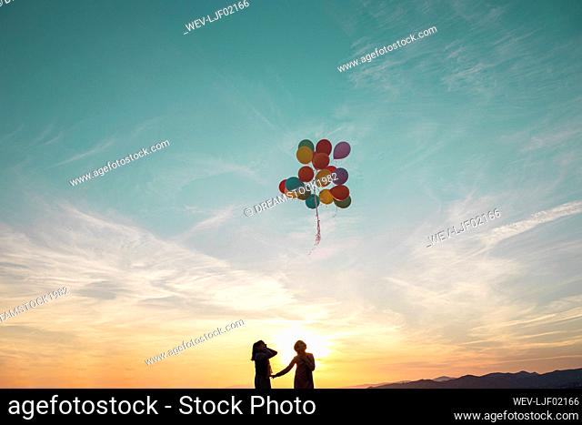 Girls in silhouette looking at helium balloons flying in air during sunset