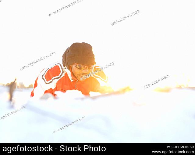 Sportsman wearing sunglasses and knit hat lying on snow during sunset