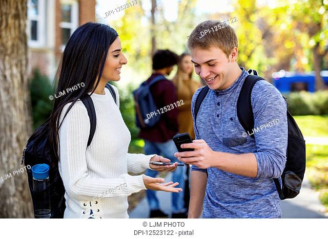 A male and female university student stand on a path looking at a smart phone together, laughing and talking together with a small group of fellow students in...